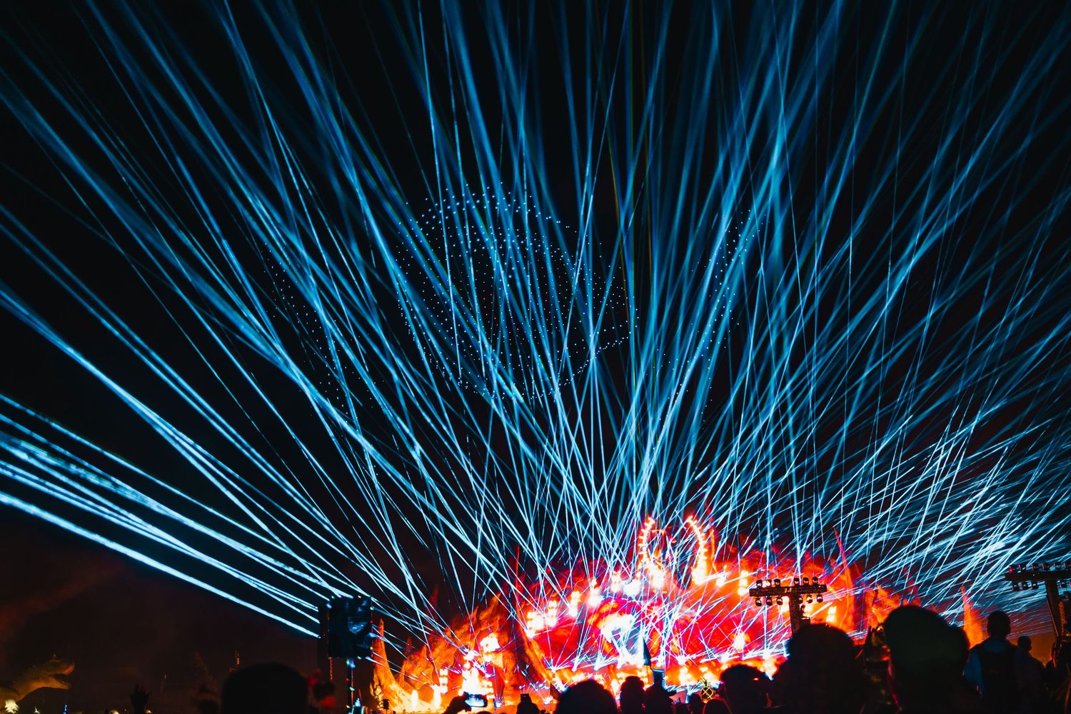 Blue laser beams shoot upward into the night sky while red flames illuminate the horizon.