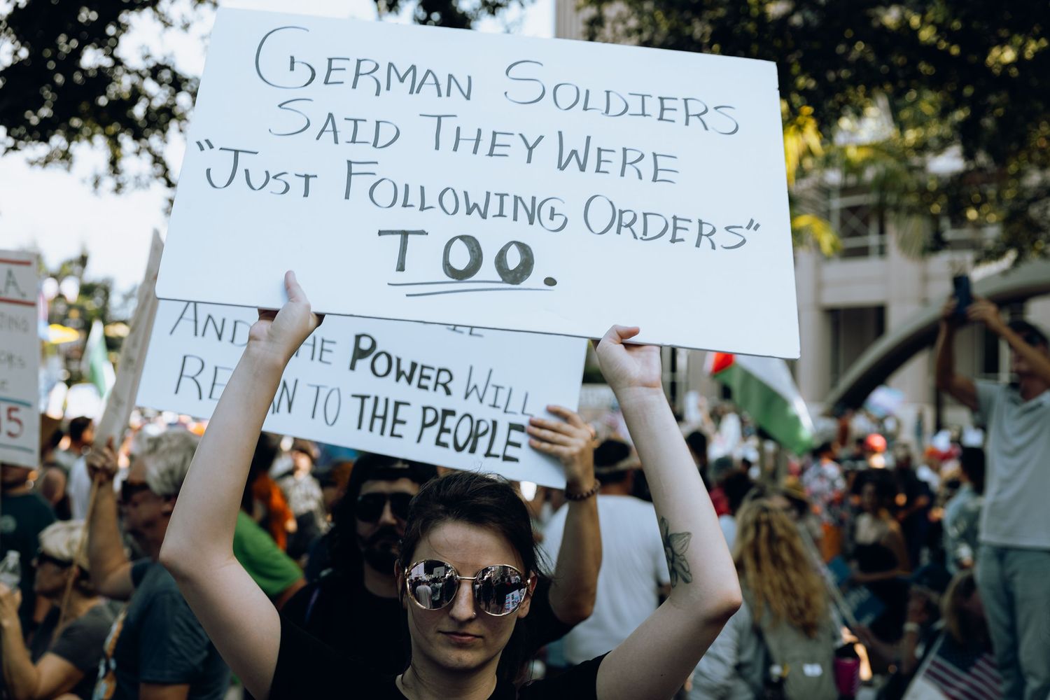 Person holding protest sign referencing German soldiers and orders among crowd of demonstrators.