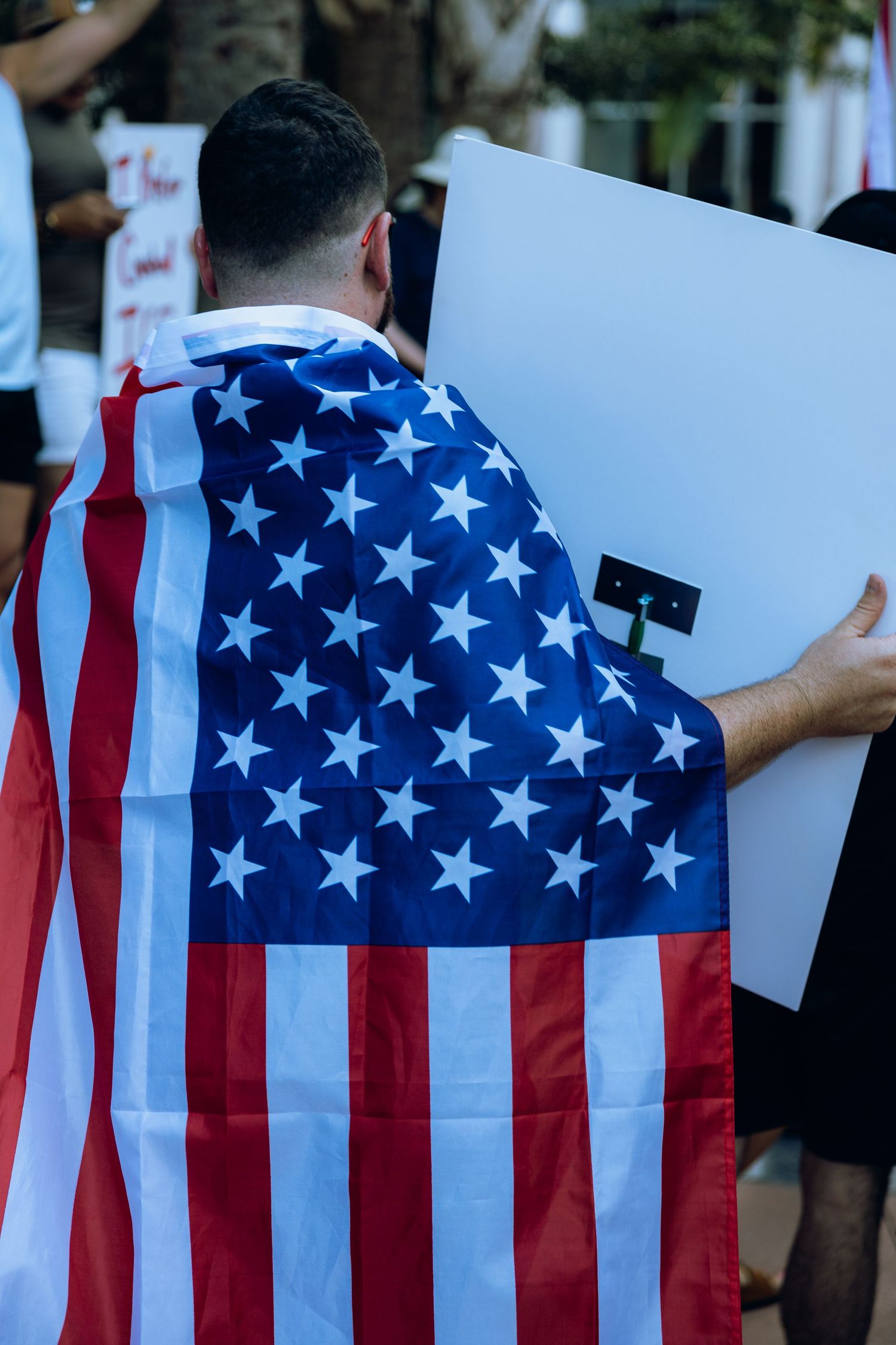 Demonstrator draped in American flag holds sign at outdoor protest.