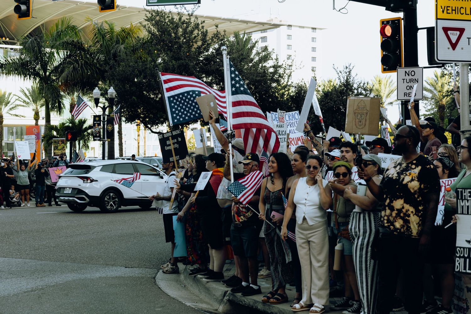 A crowd of demonstrators wave American flags and banners while gathering along a palm tree-lined street corner.