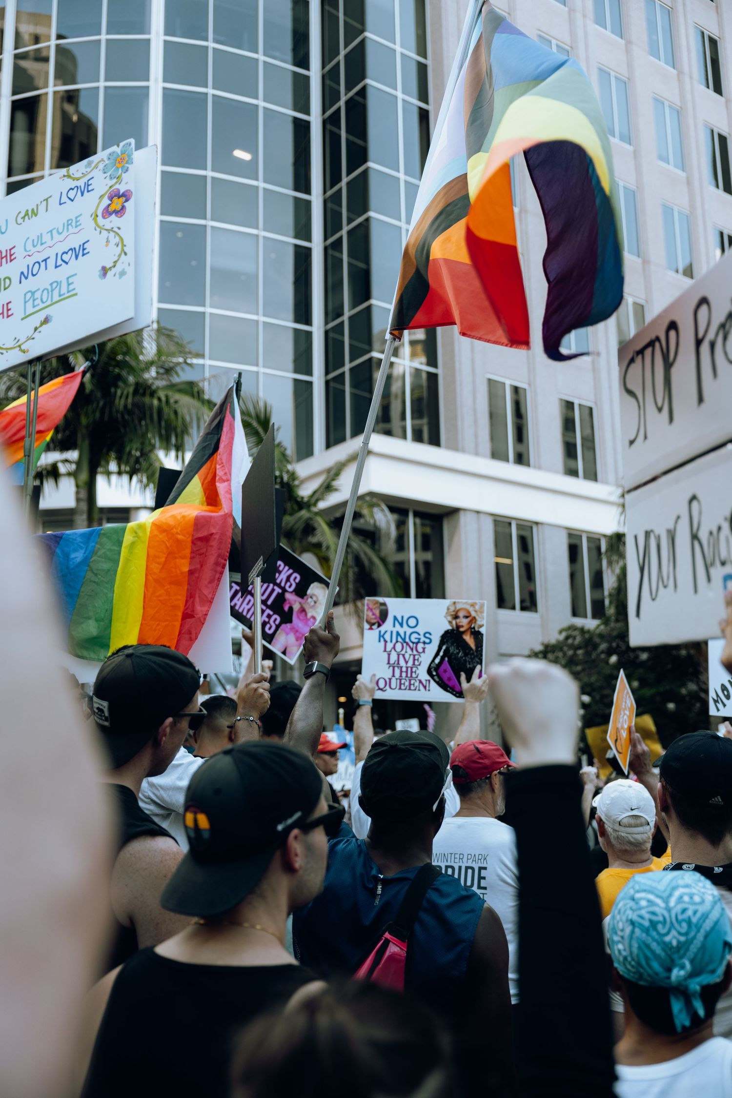 Colorful pride flags flying over crowd of protesters near urban building.