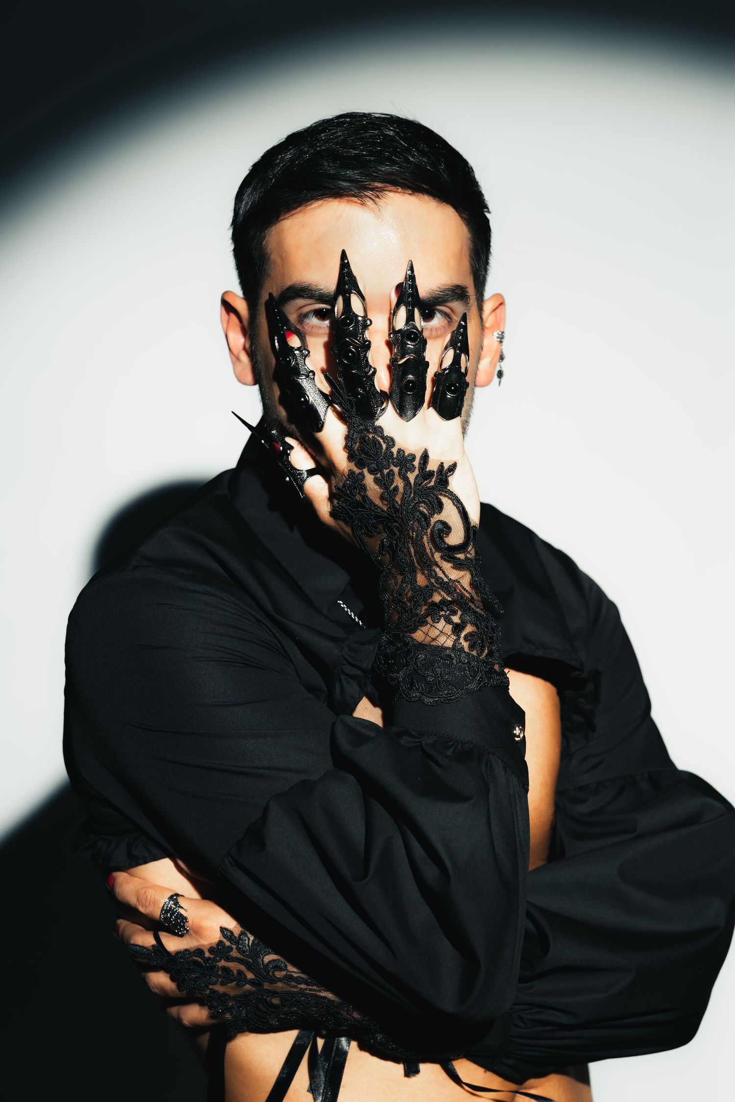 Man in black shirt holding ornate black lace glove over face in dramatic white light.