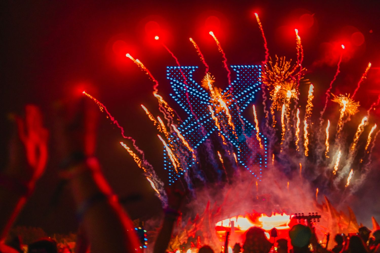 Spectacular fireworks display over a music festival stage with red and blue lights illuminating the crowd.