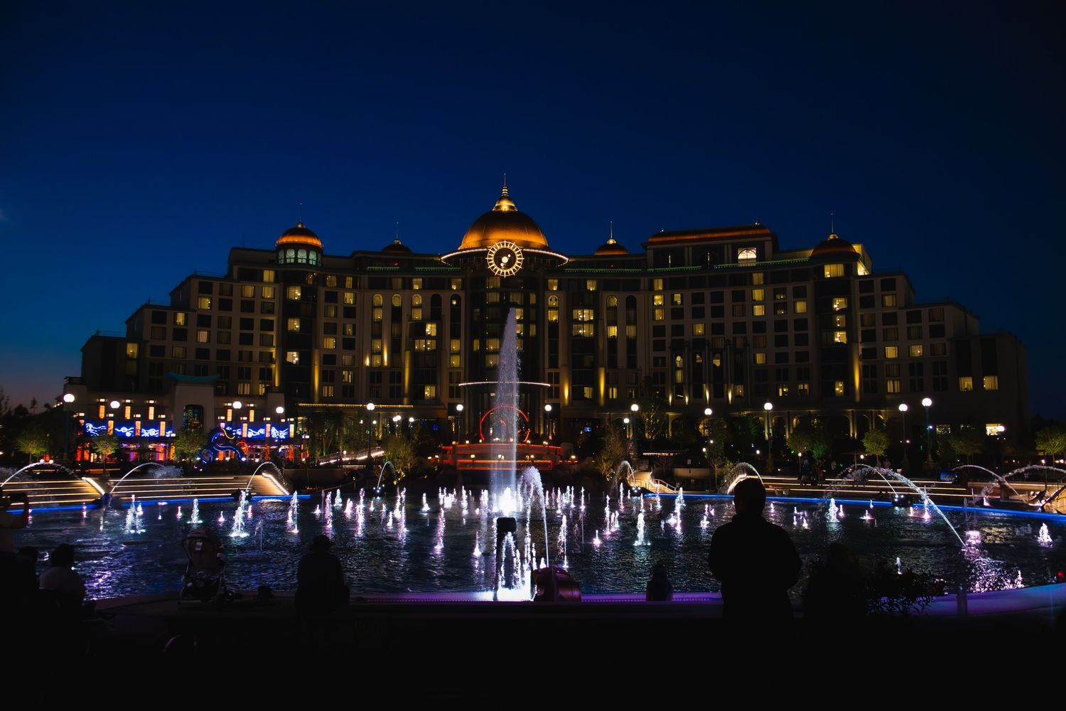 Illuminated hotel building at night with colorful fountain display in front creating a magical waterfront ambiance.