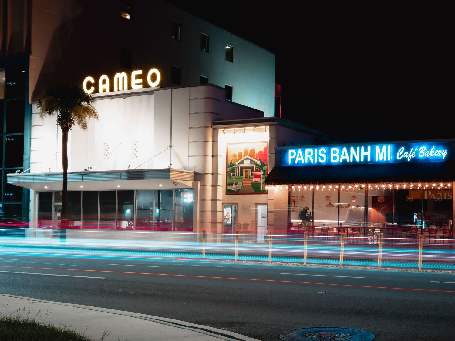 Long exposure night shot of the illuminated Cameo theater and Paris Banh Mi storefront with light trails on the street.
