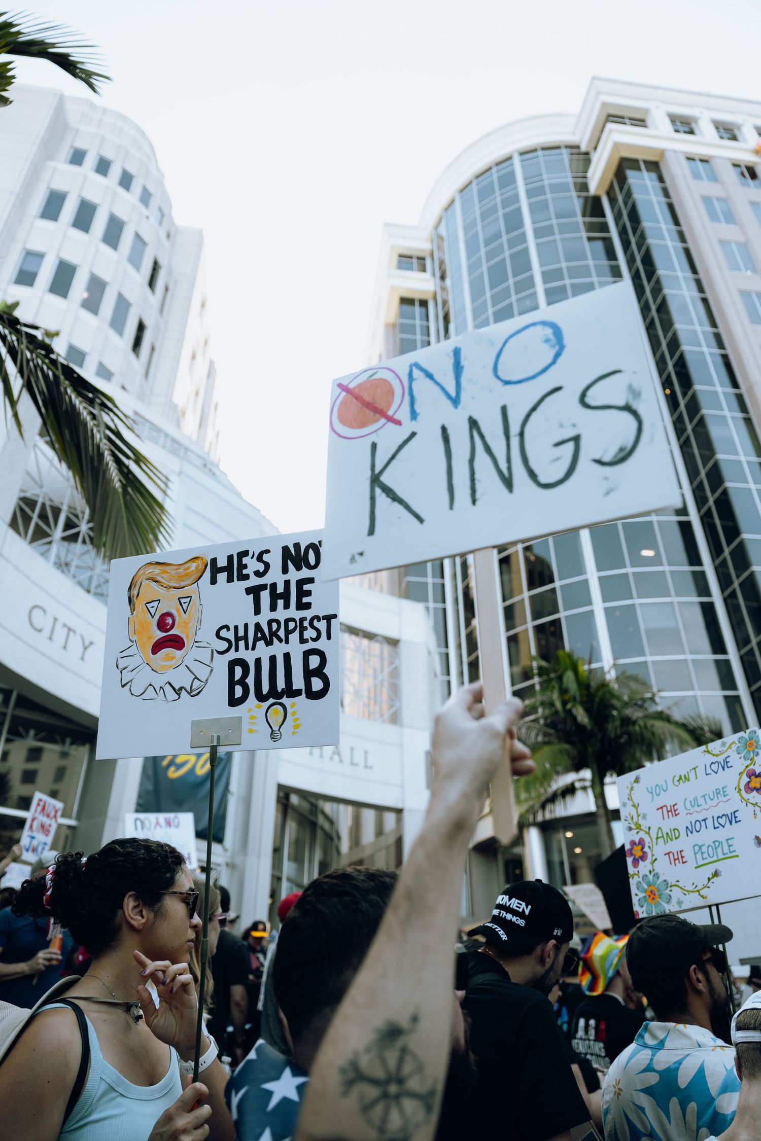 Crowd of protesters holding signs outside a modern glass office building with palm trees.