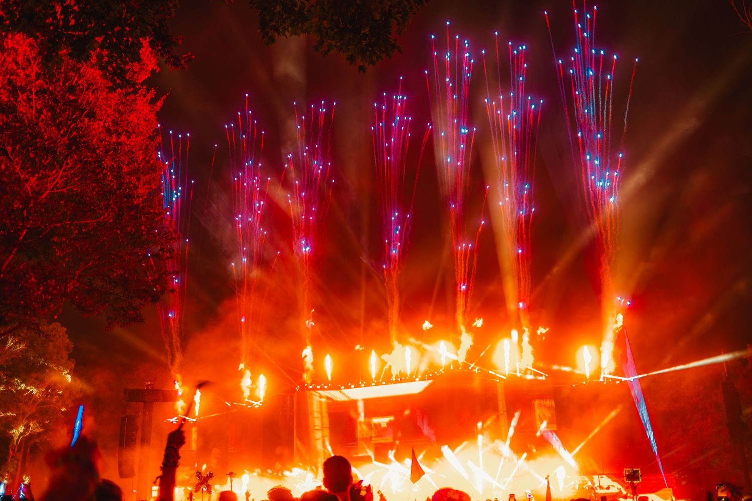 Dramatic outdoor concert stage with vibrant red and purple pyrotechnics lighting up the night sky above glowing flames.