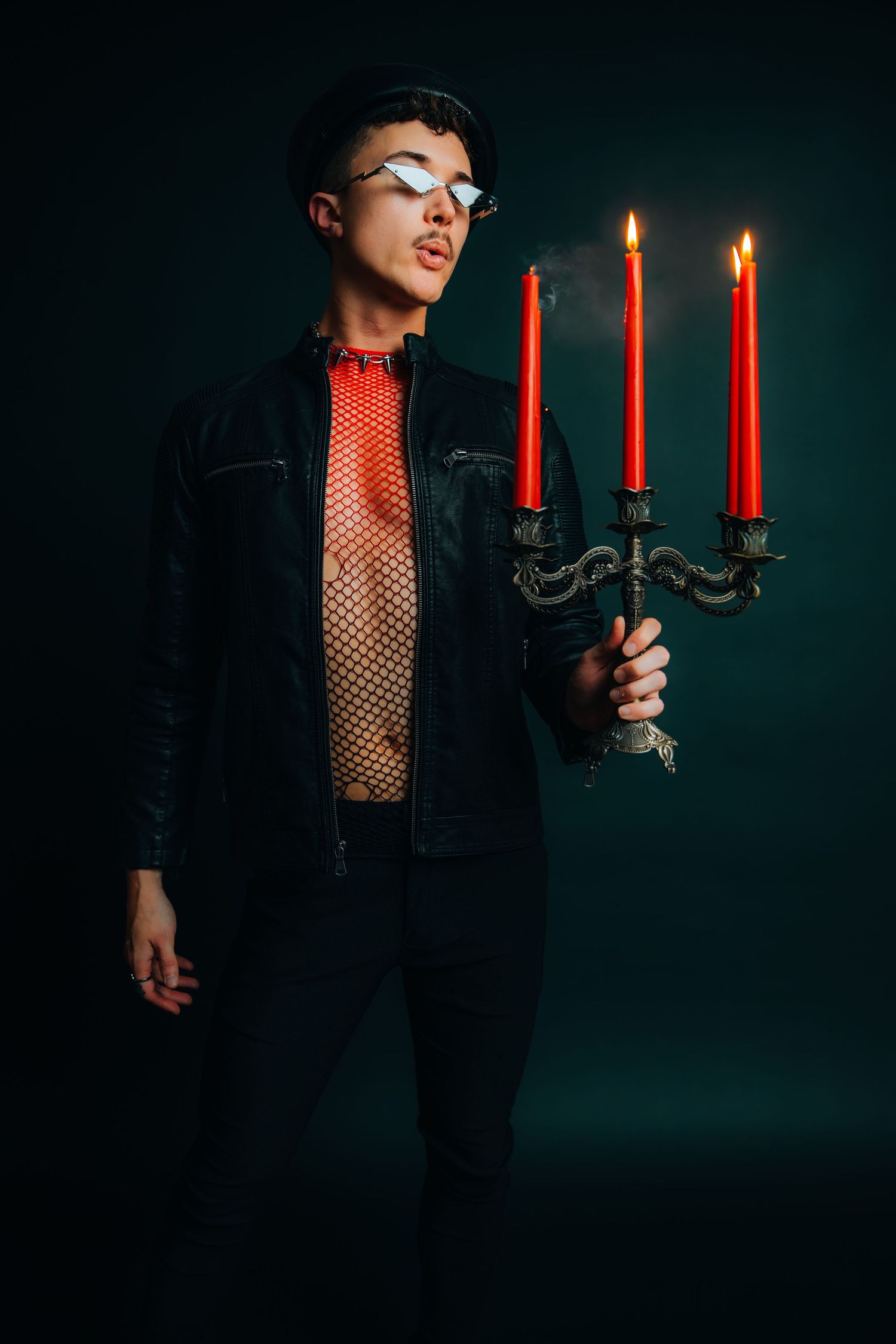 A stylish person in a black jacket and mesh top holds a candelabra with three lit red candles in a dark studio.