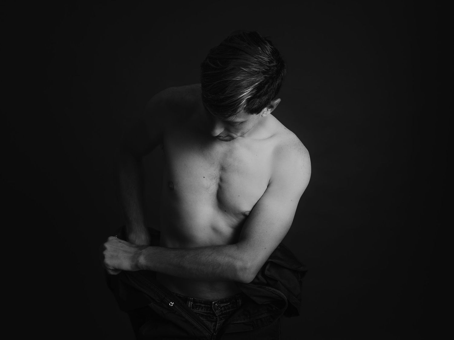 Black and white photo of a shirtless young man looking down, partially removing his shirt against a dark background.
