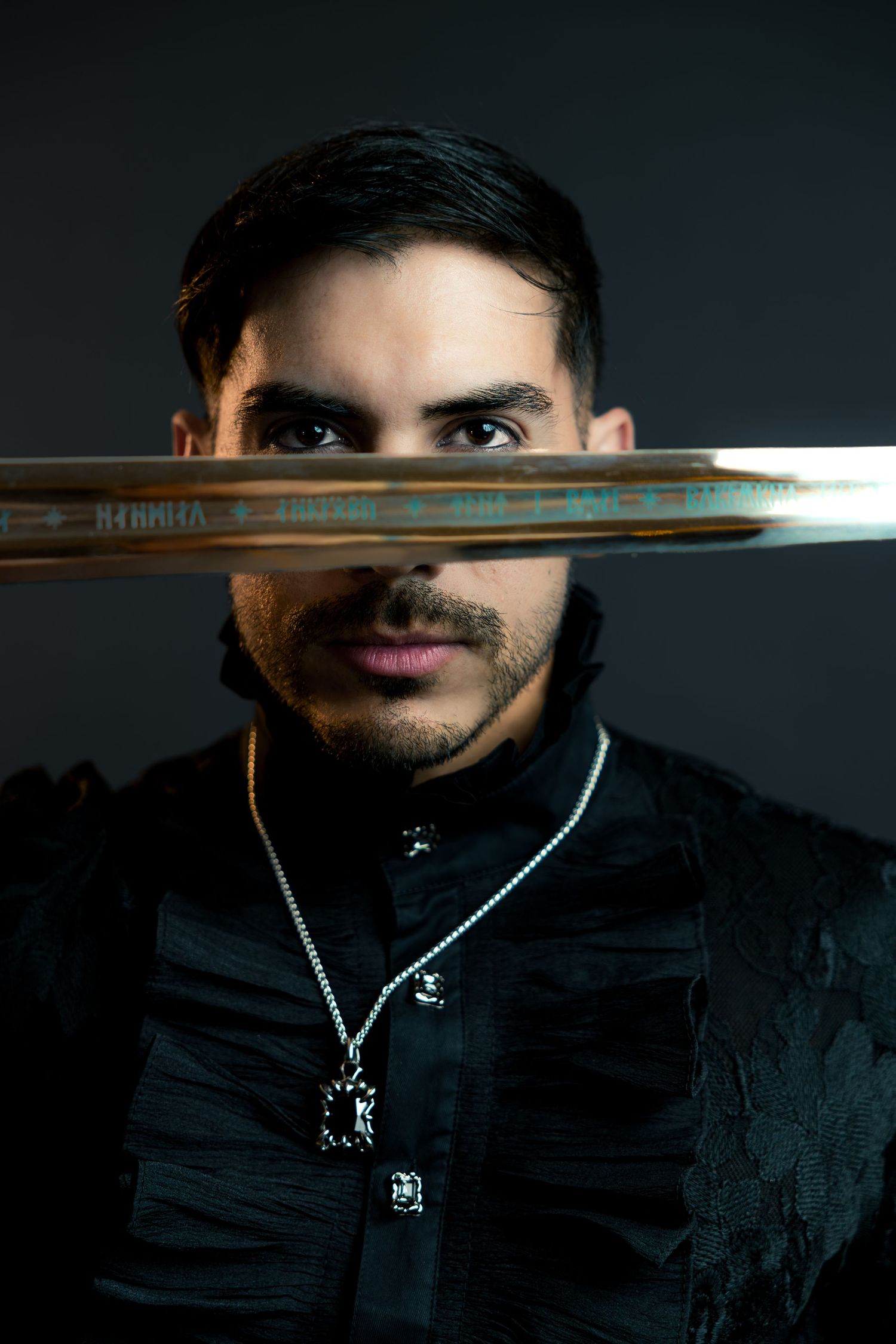 Man in black jacket holding golden sword horizontally across face with chain necklace.