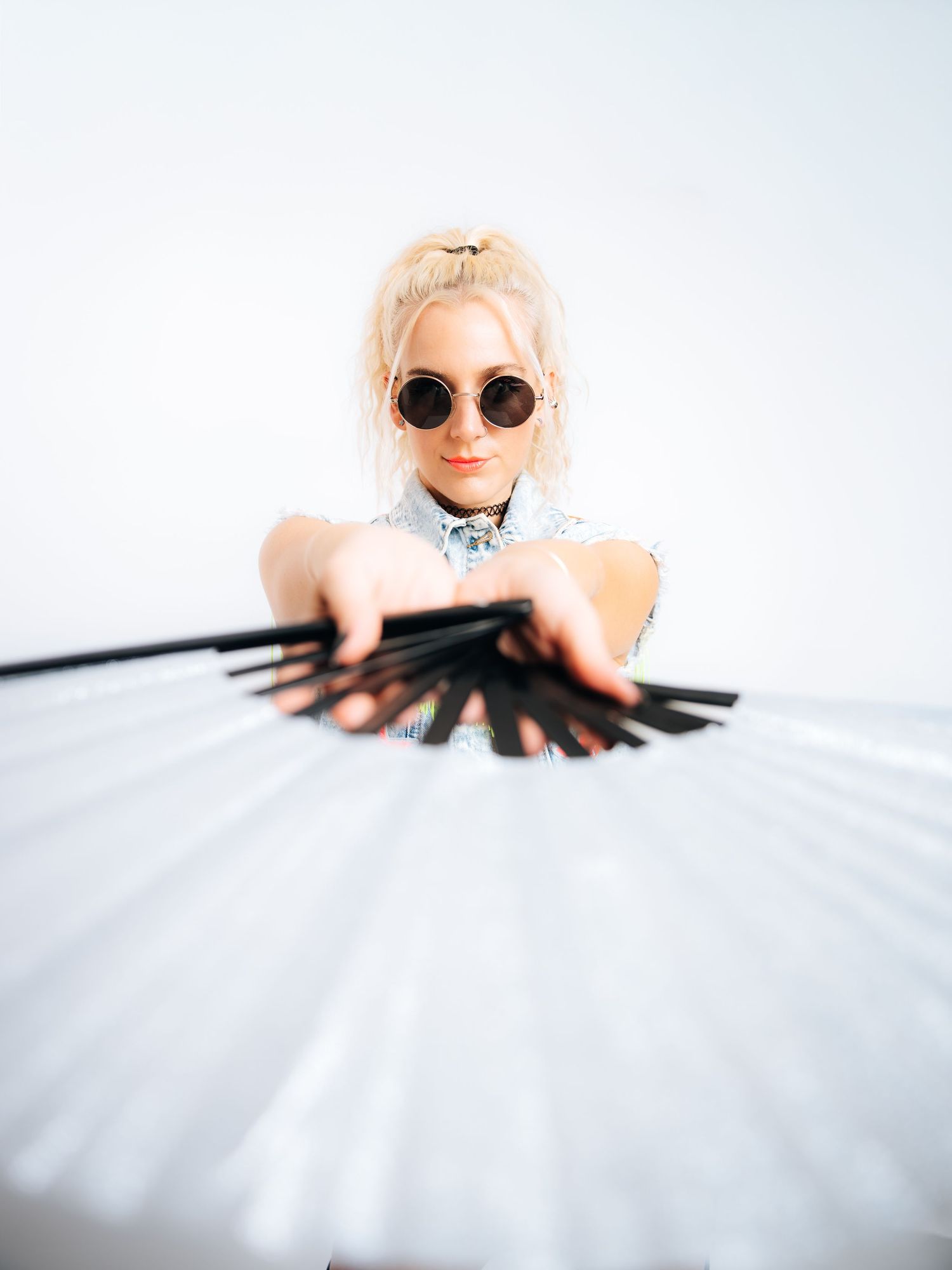 Blonde woman in round sunglasses holding a large black and white fan against a white background.
