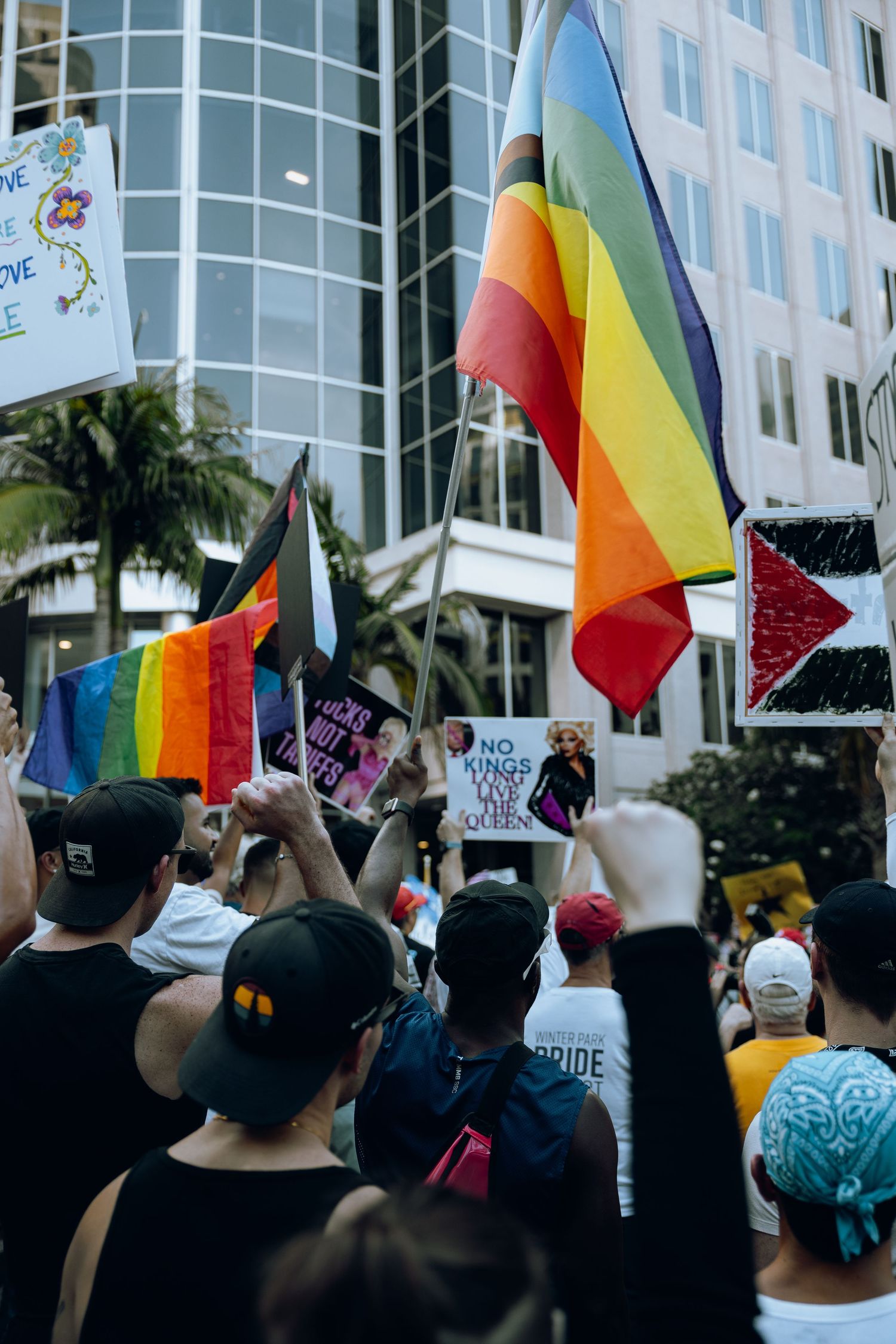 Rainbow flags wave above crowd of protesters outside modern office building.