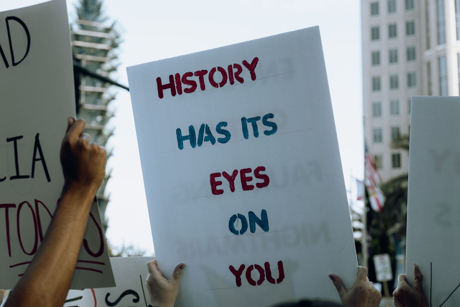 A protest sign reads 'History Has Its Eyes On You' against an urban backdrop.