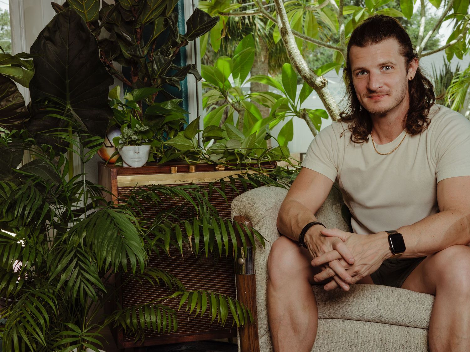 Person relaxes on a beige couch surrounded by lush green houseplants in a bright indoor setting.