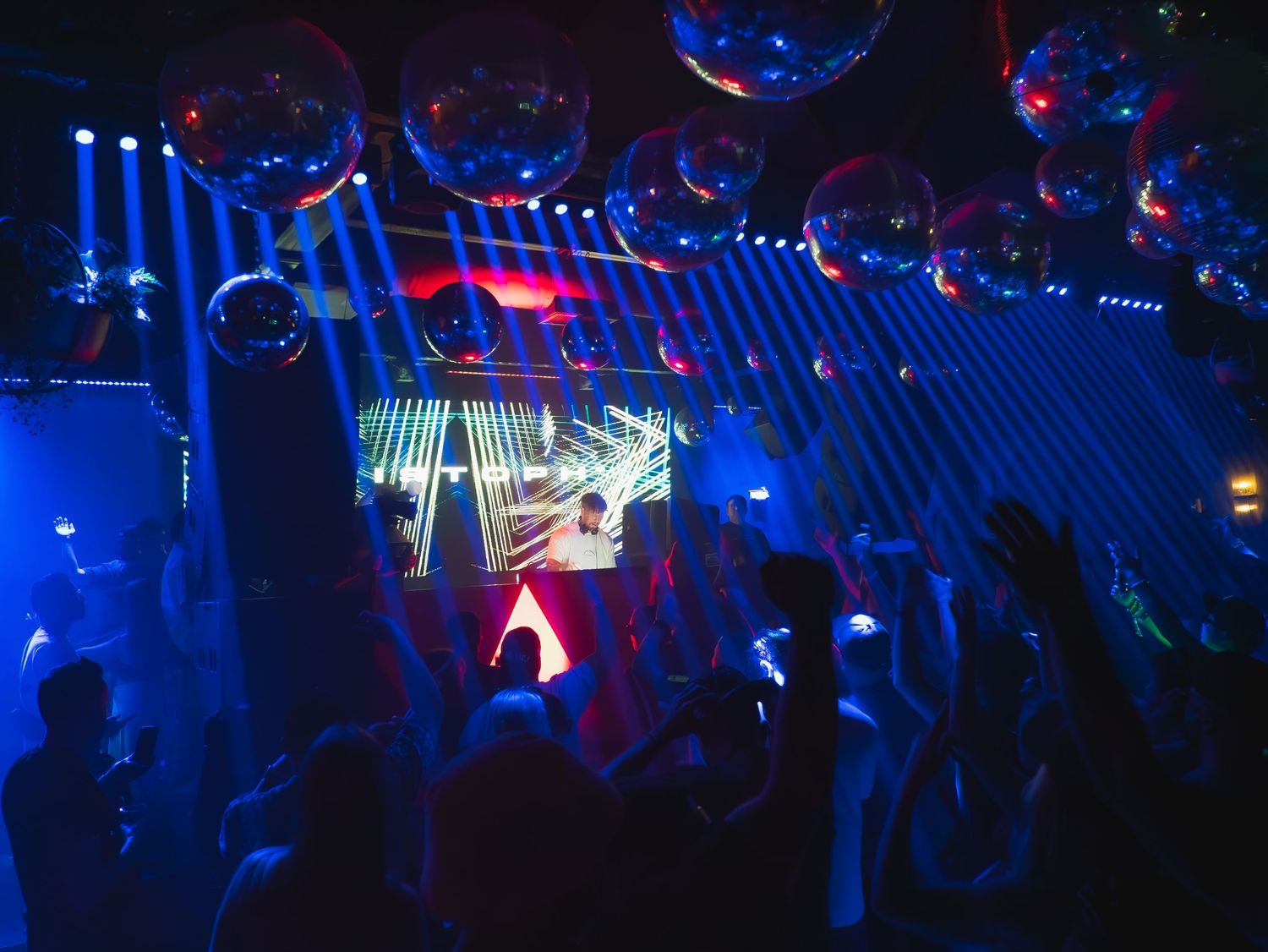 A nightclub interior with blue lighting, disco balls overhead, and silhouetted crowd enjoying the dance floor atmosphere.