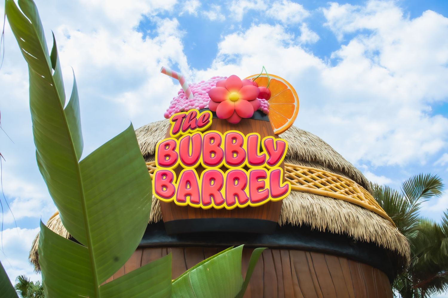 A tiki-style Bubbly Barrel restaurant sign with pink neon letters against a blue sky with palm trees and thatched roof.