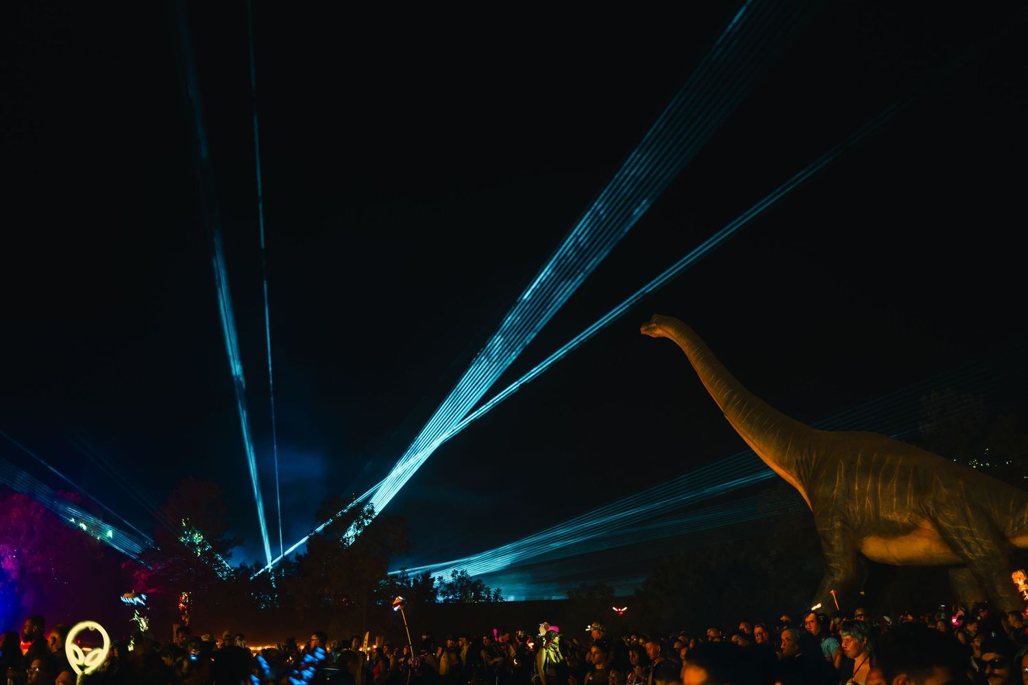 Blue laser beams illuminate the night sky during an outdoor festival with glowing lights and silhouetted crowd.
