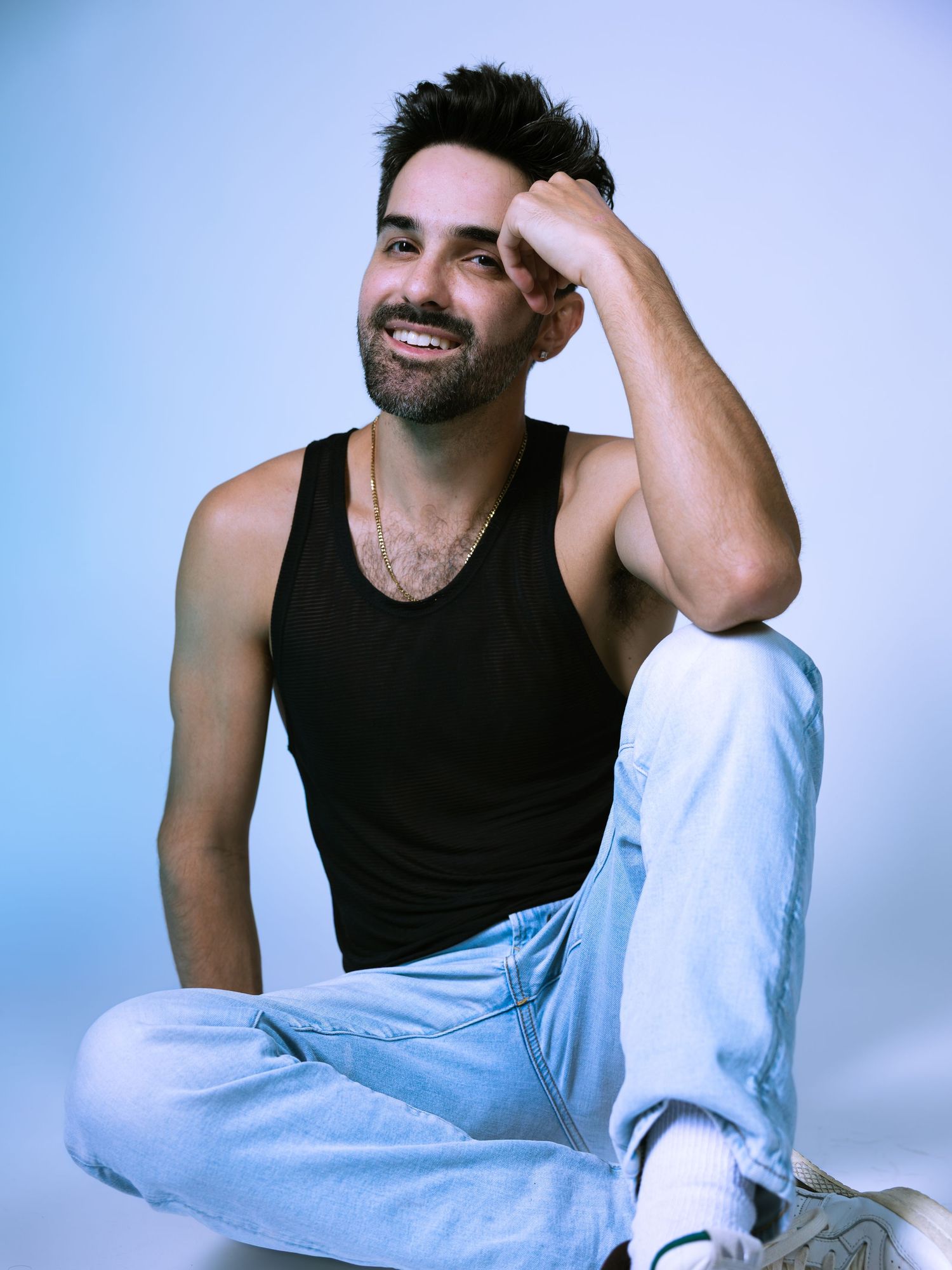 Smiling man in black tank top and light blue jeans sitting cross-legged on floor against blue background.