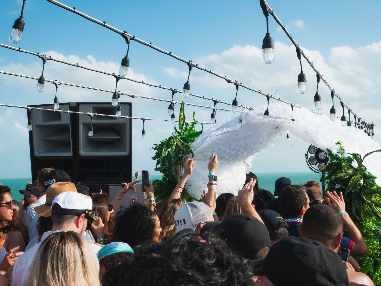 Crowd dancing at an outdoor party with string lights overhead, speakers, and ocean views in the background.