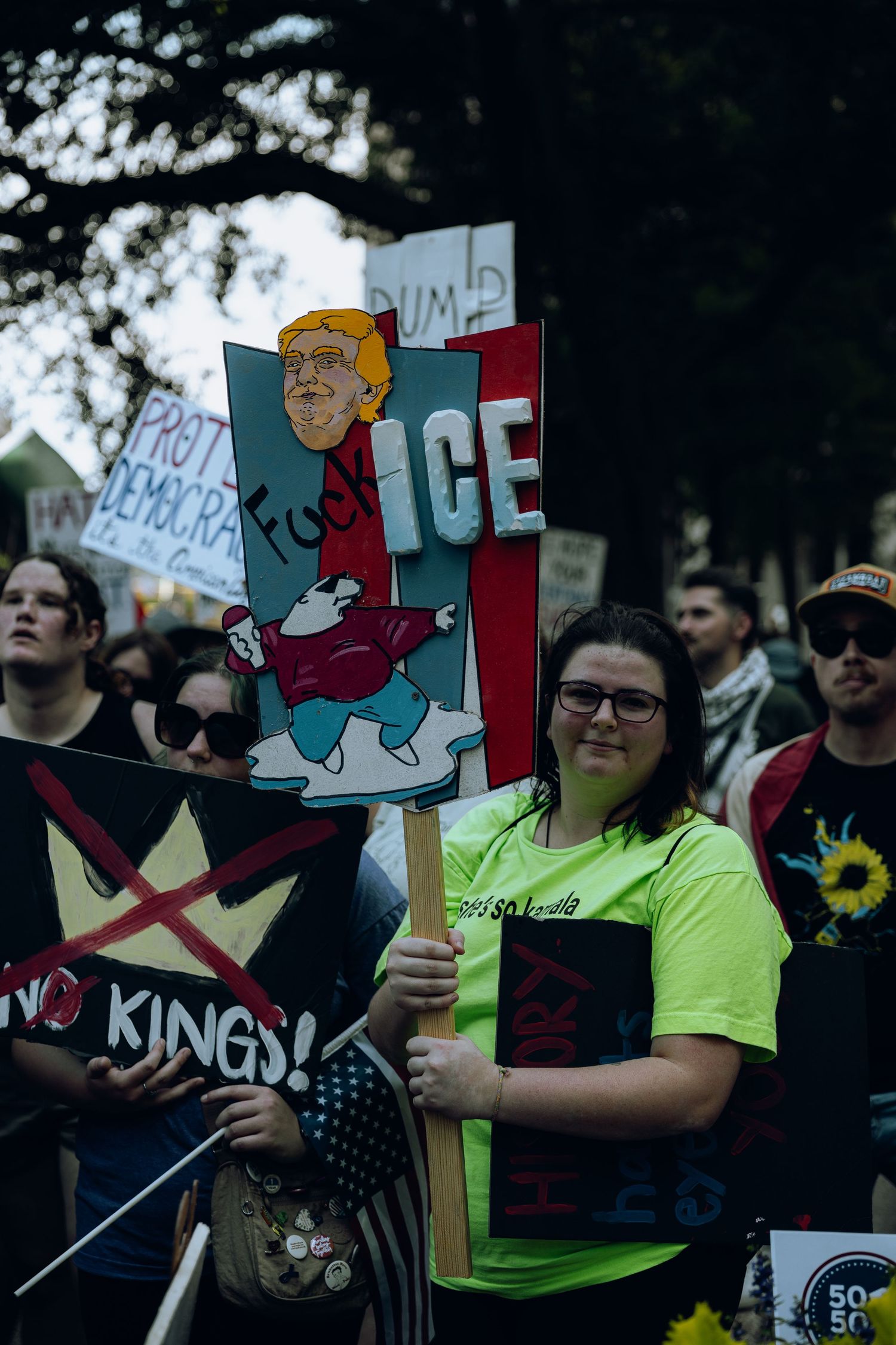 Protesters hold signs at a political demonstration with one prominent sign featuring an ICE-themed political cartoon.