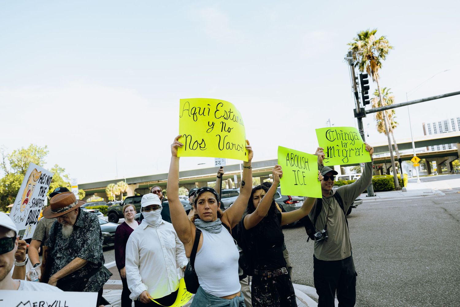 Group of protesters holding bright neon yellow signs gather outside on a sunny day with palm trees in background.