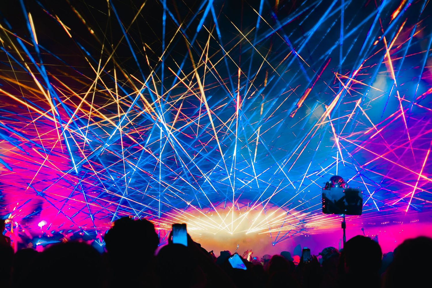 Vibrant laser light show at a music festival with colorful beams of pink, blue, and yellow over a crowd.