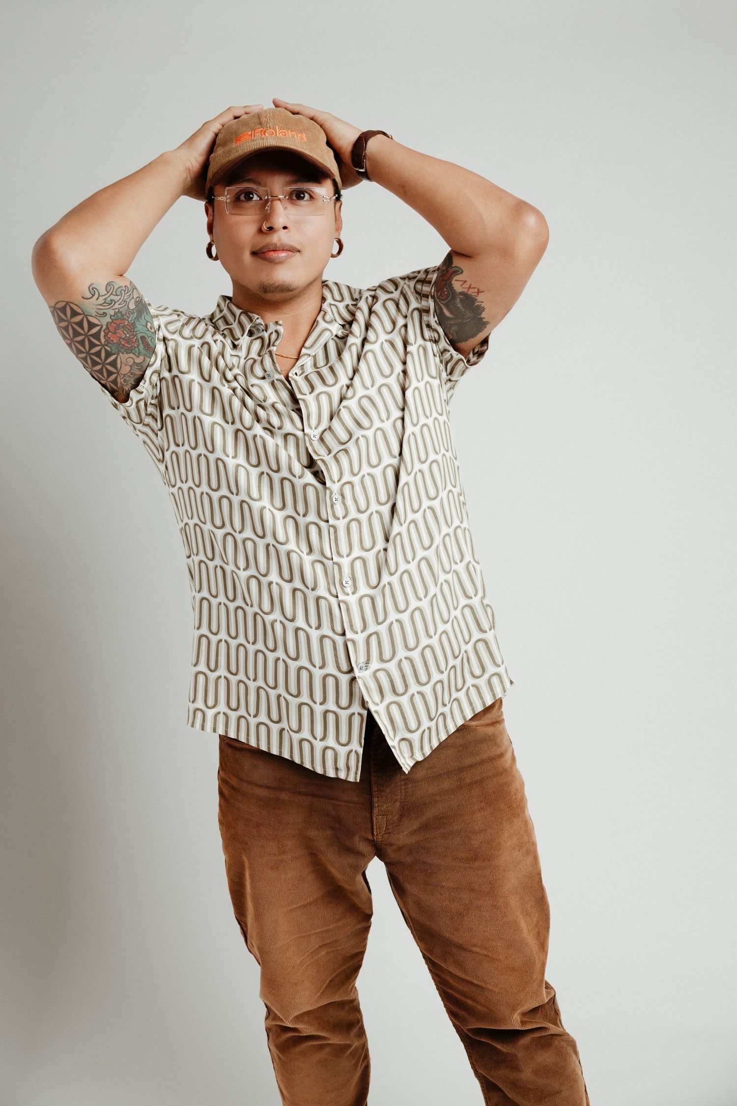 Tattooed man in patterned shirt, brown cap and pants poses with hands on head against white background.