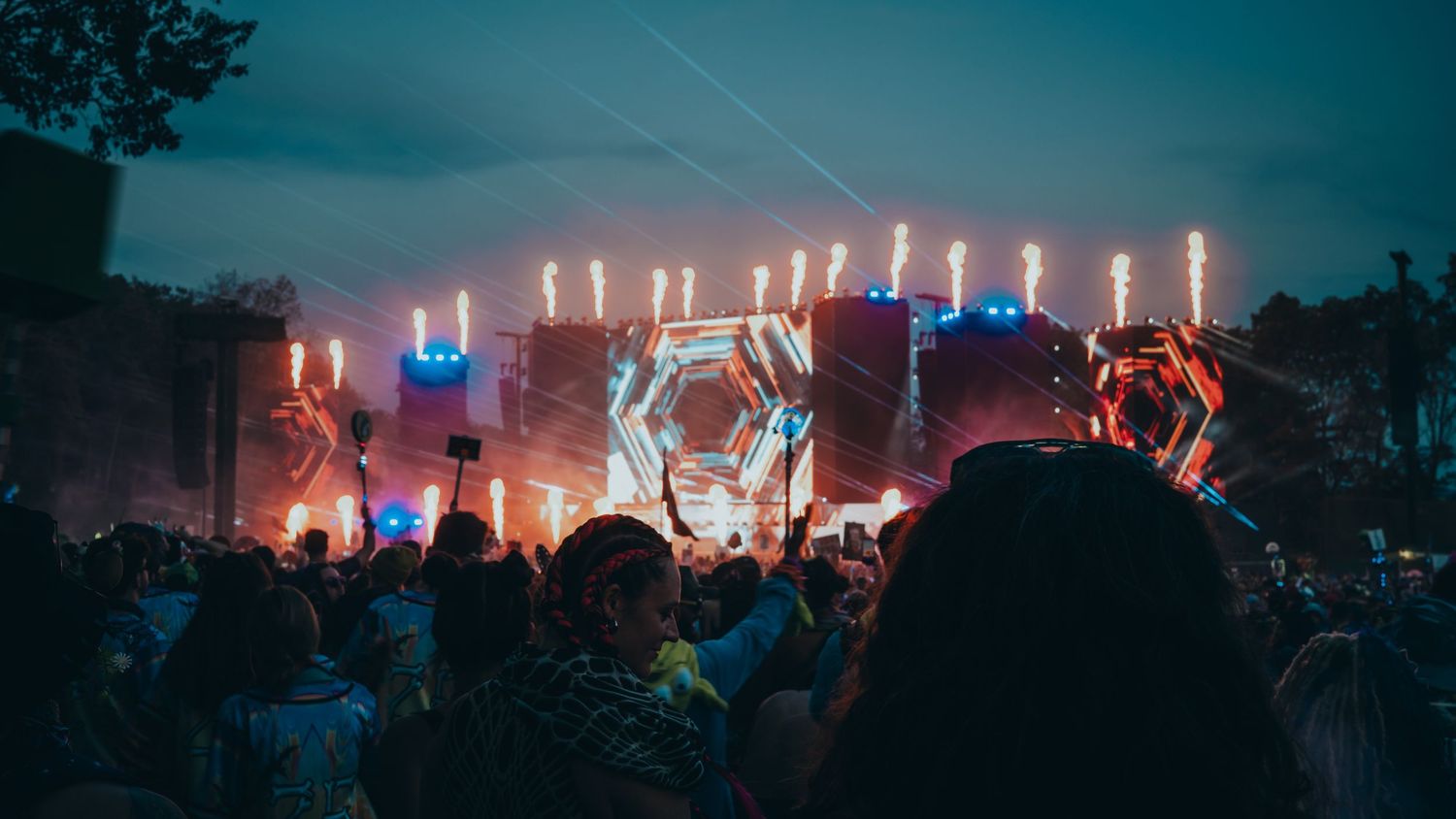 Crowd at outdoor music festival with colorful stage lights and fireworks lighting up the night sky.