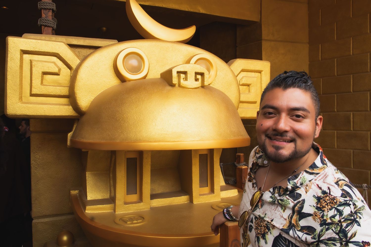 A smiling visitor poses next to a large golden cartoon character sculpture mounted on a brick wall.
