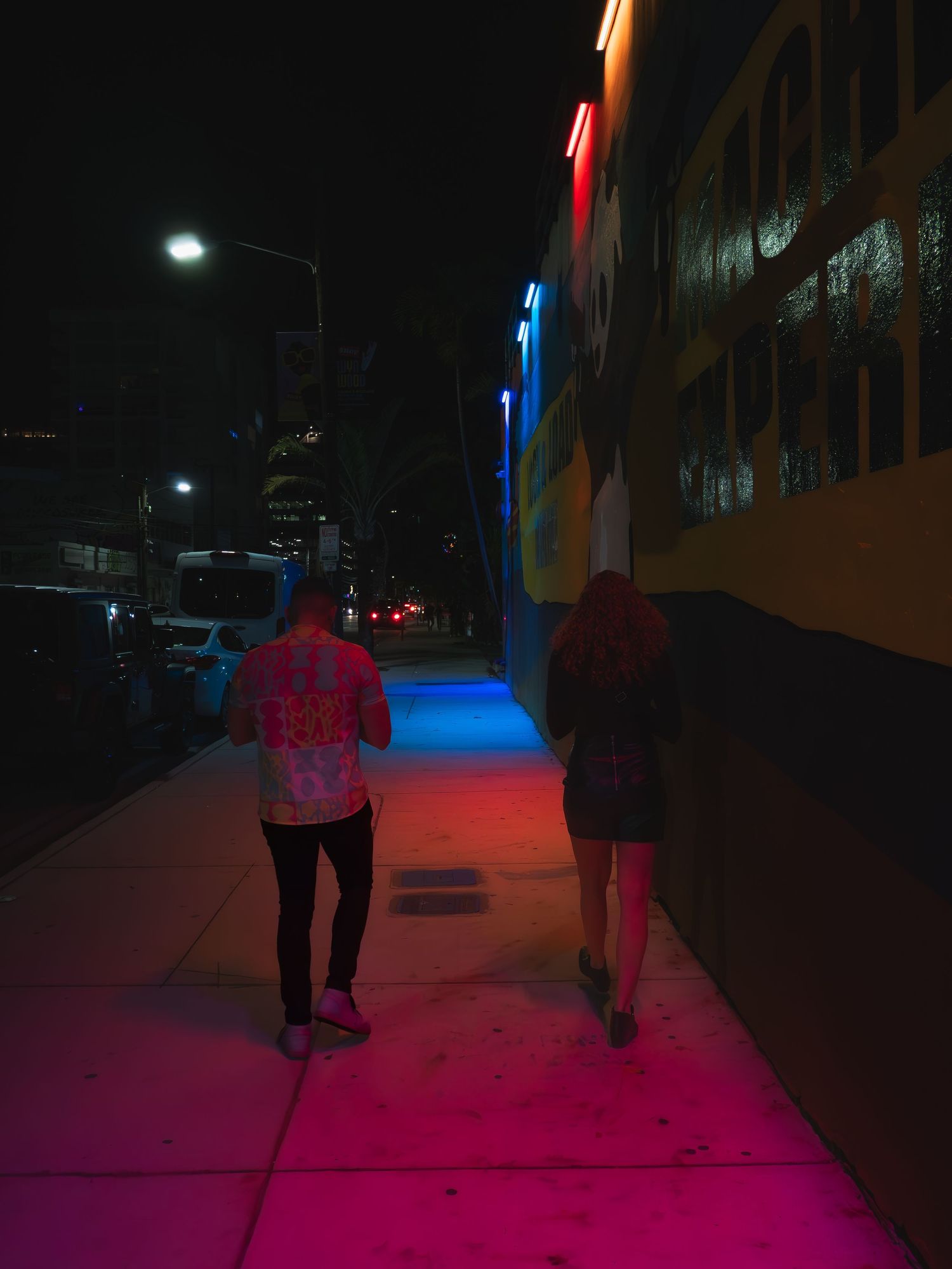 Two people walking at night on a sidewalk illuminated by vibrant red and blue neon lights from nearby buildings.