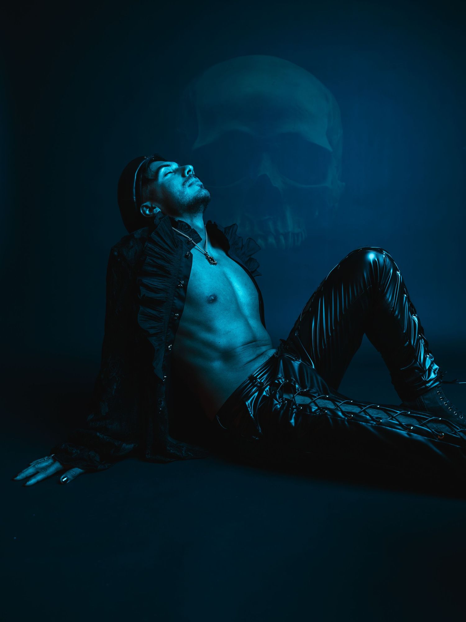 A shirtless man in black leather pants reclines dramatically against a dark blue backdrop with a glowing skull silhouette.