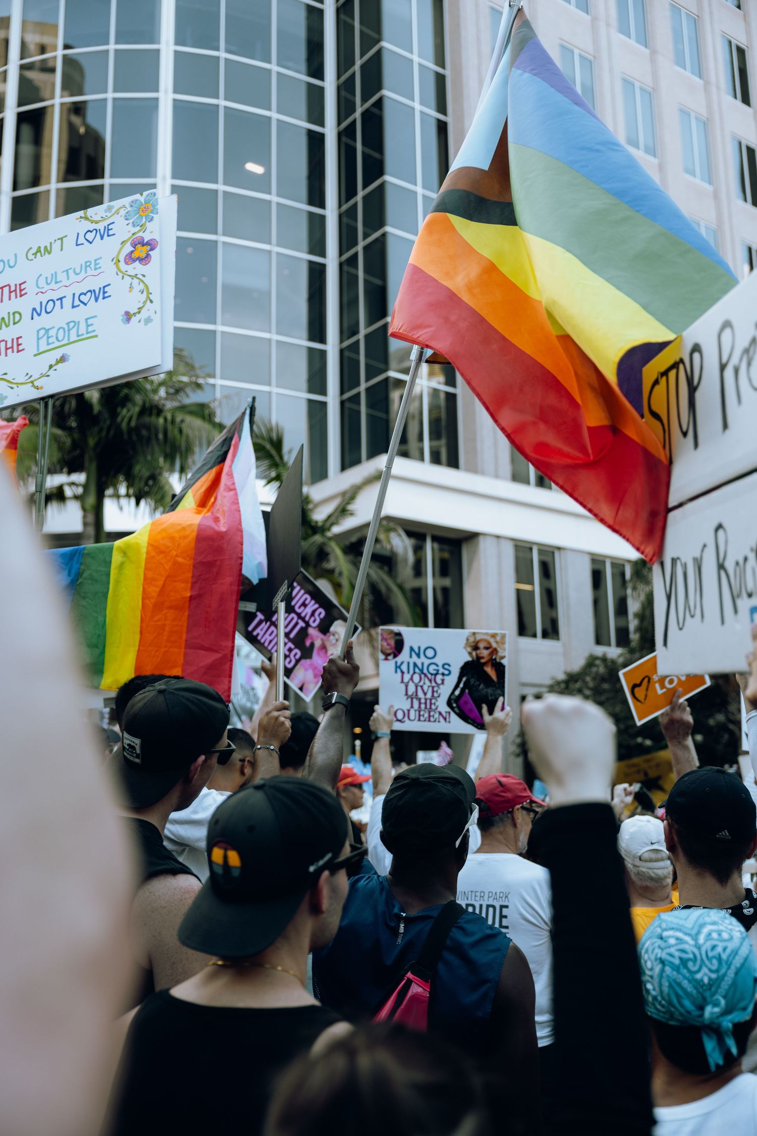 Rainbow flags and protest signs fill the scene as demonstrators rally outside a modern office building.