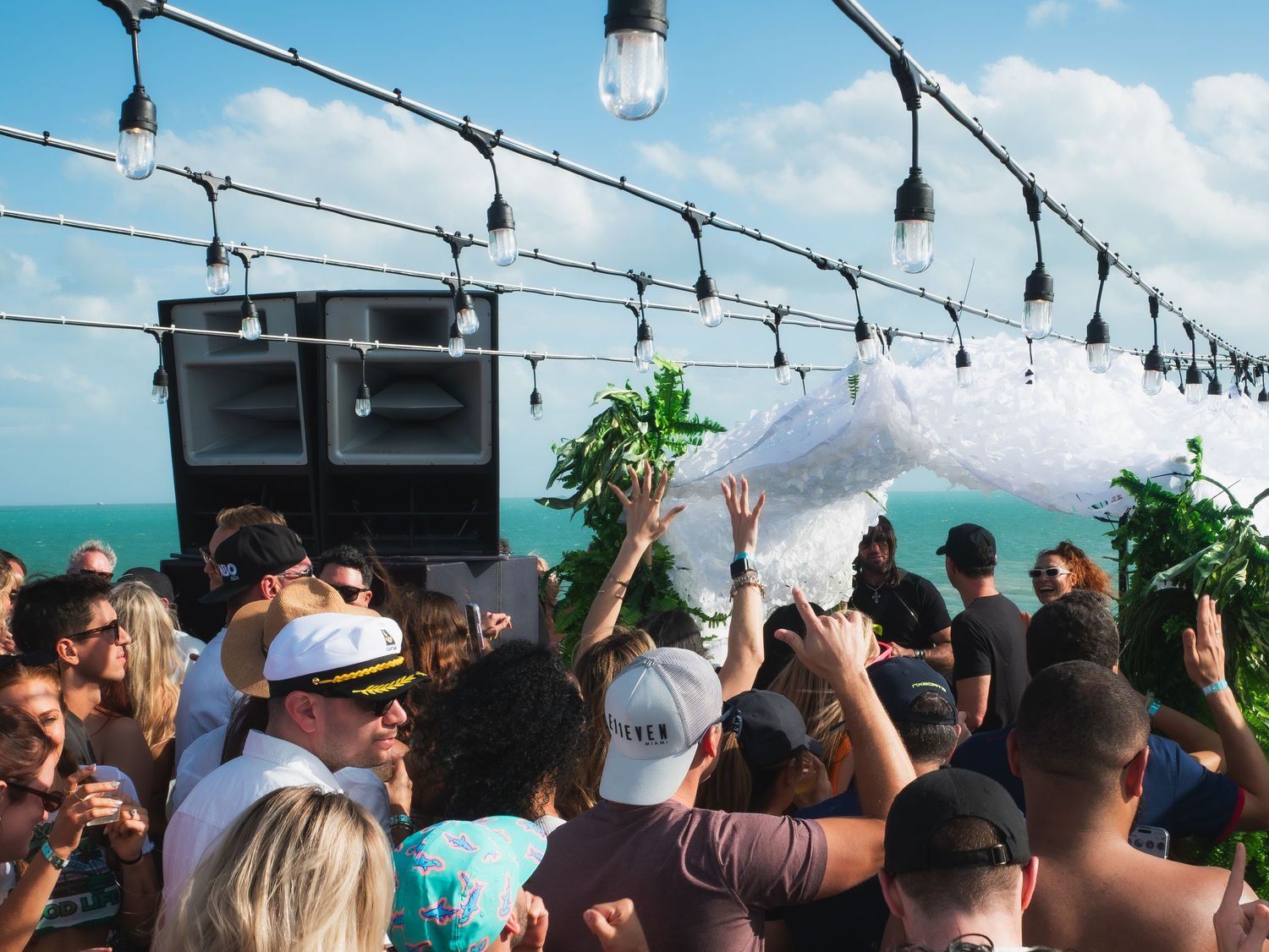 Crowd dancing at a rooftop party with string lights and ocean views during a daytime celebration.