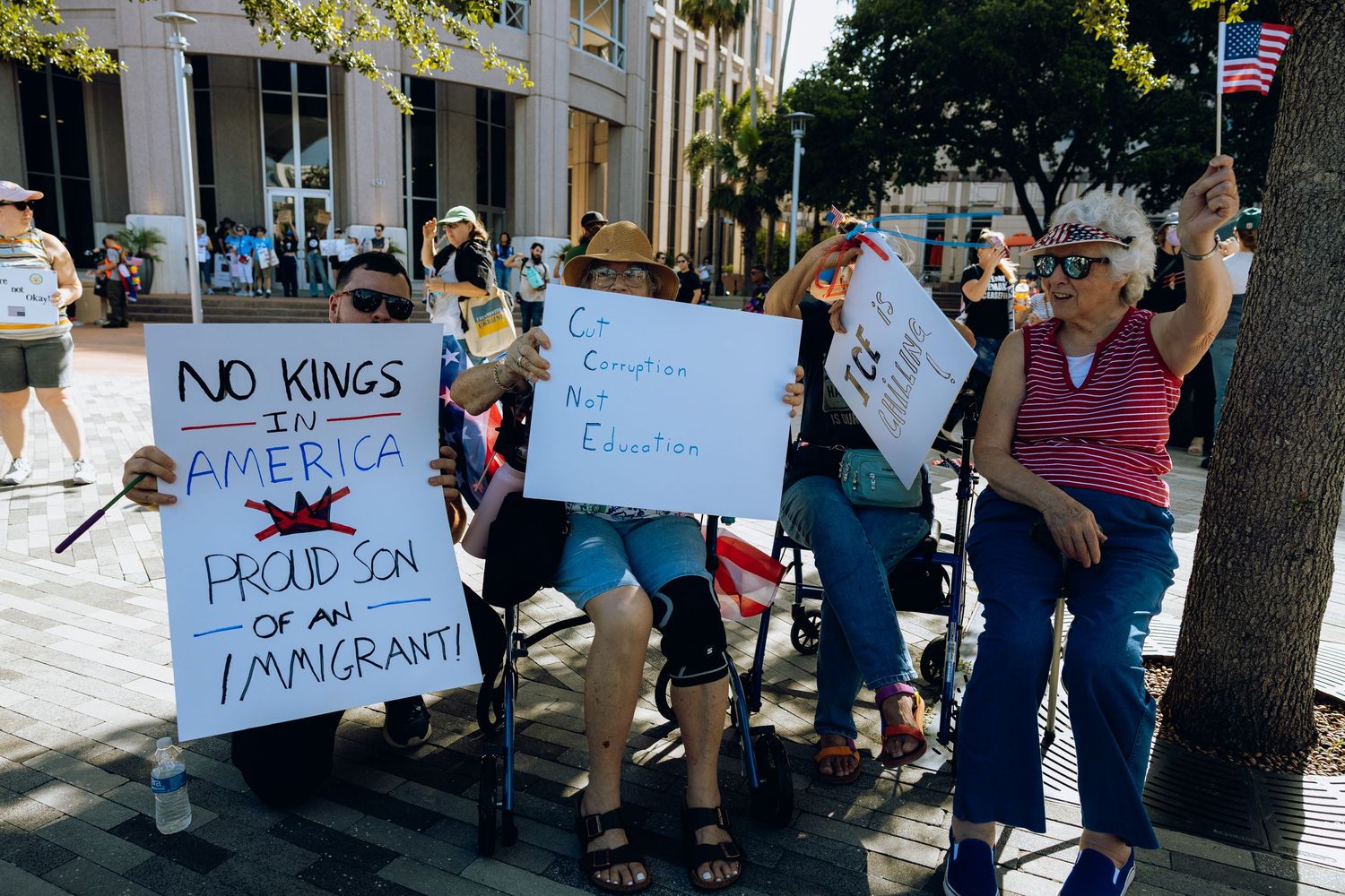 People gather on a sidewalk holding protest signs with an American flag visible in the background.