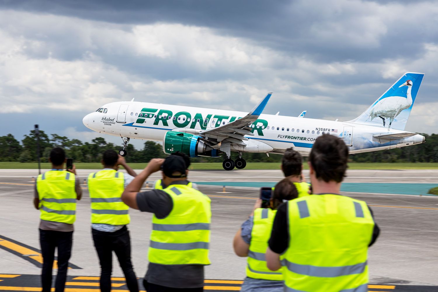 Frontier Airlines Airbus A320 aircraft taxiing on runway while airport ground crew in yellow vests watch and photograph.