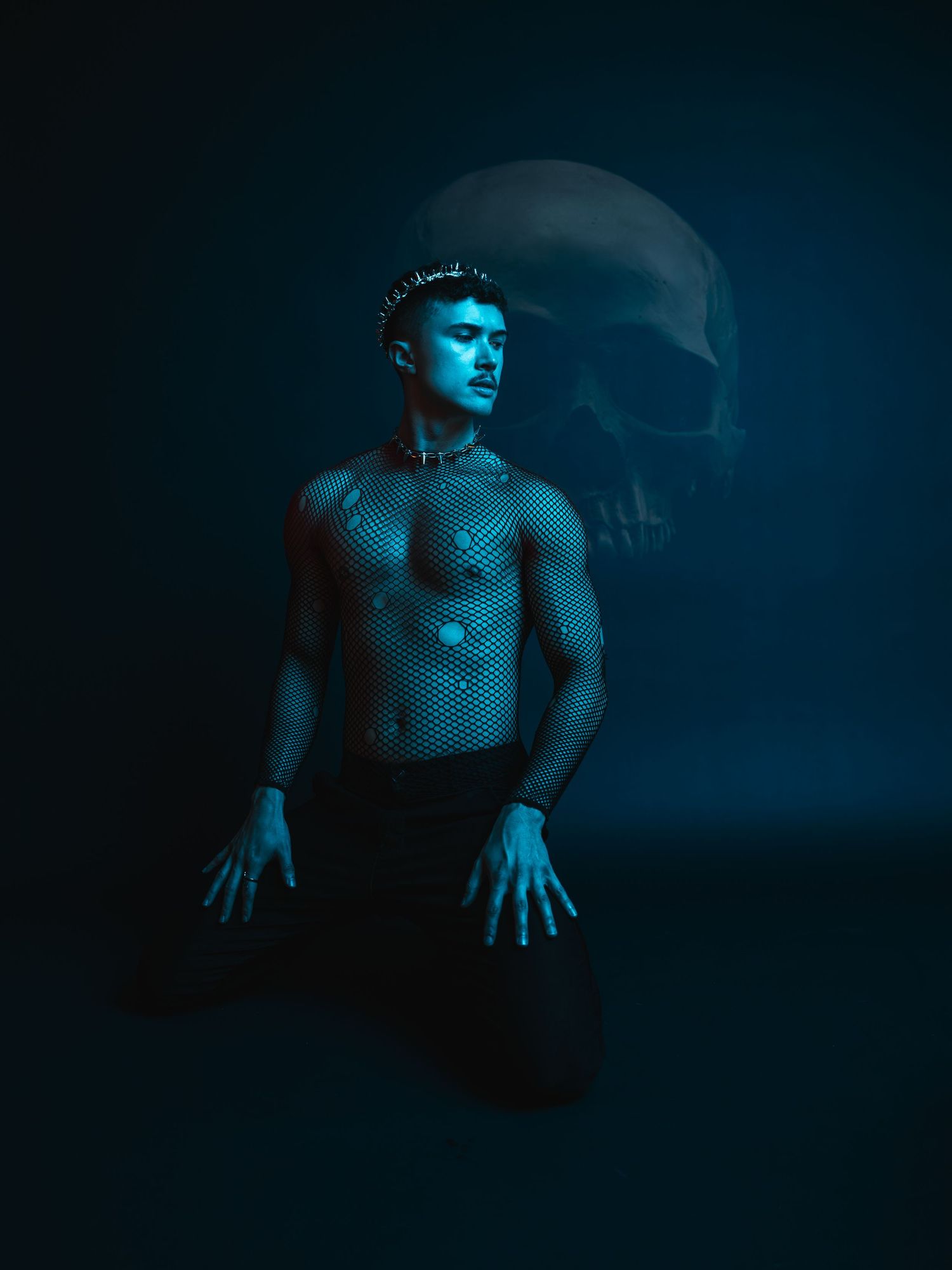 Shirtless man in mesh top kneeling dramatically under blue light with large skull looming in dark background.