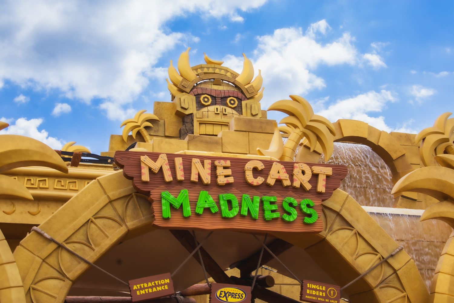 The entrance sign for Mine Cart Madness attraction features golden archway decorations against a bright blue sky.