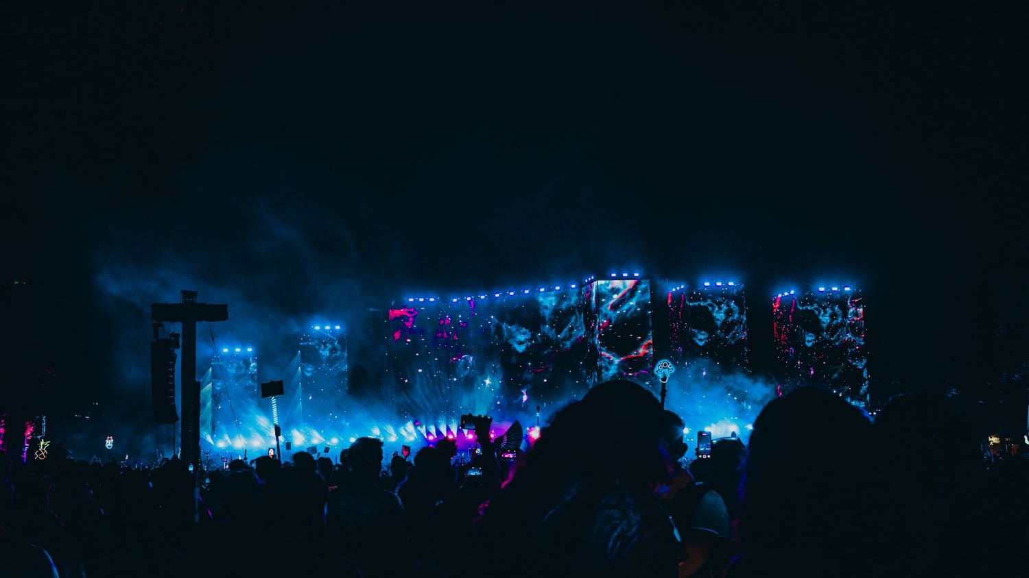 Concert crowd silhouettes against a dark sky with vibrant blue and purple stage lighting creating a dramatic nighttime atmosphere.