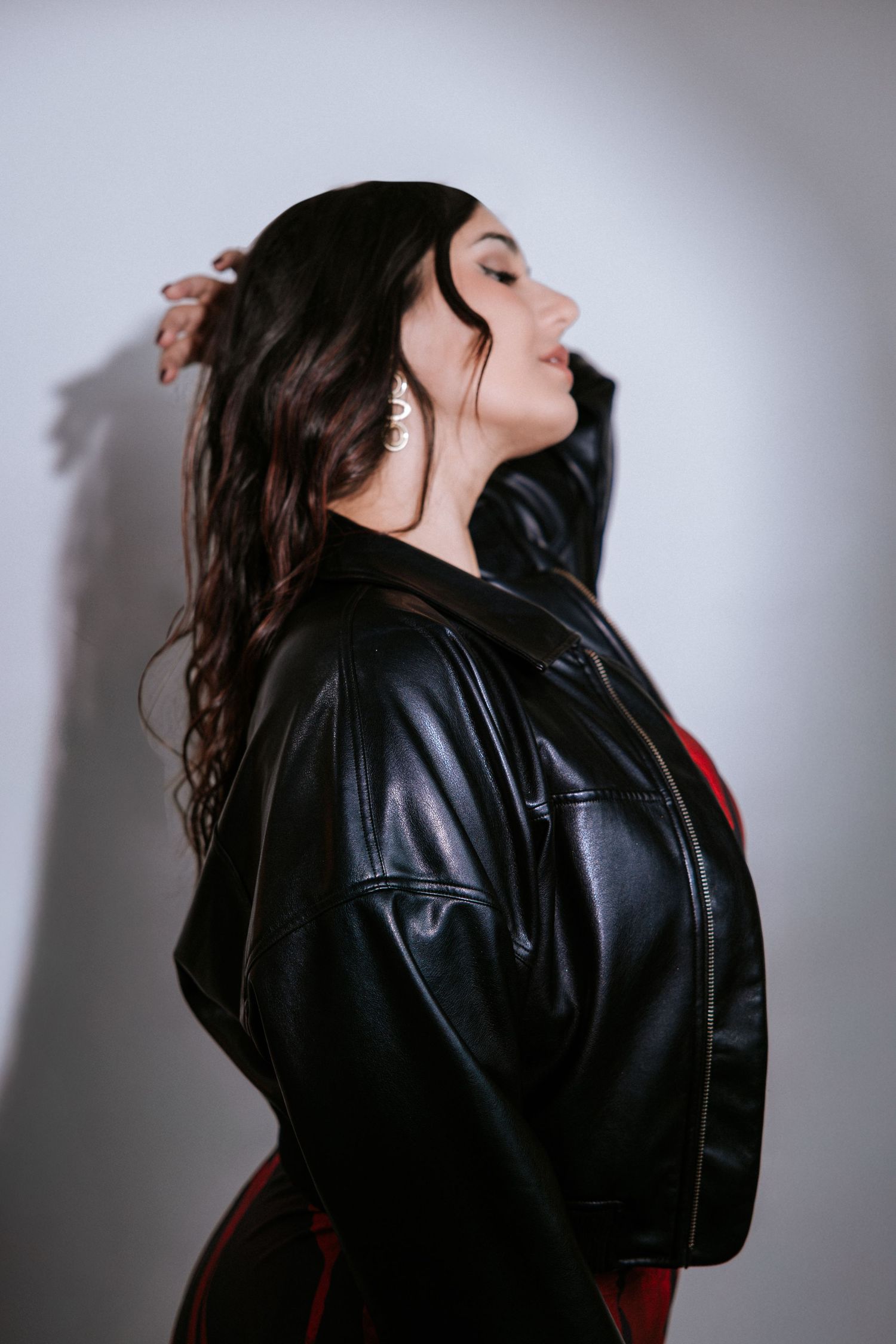 Woman in black leather jacket posing sideways with eyes closed against white wall.