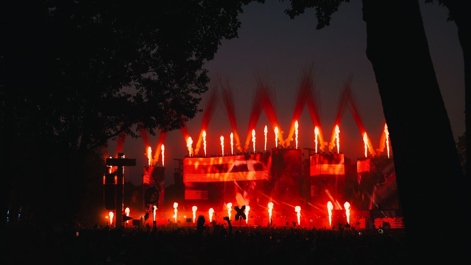 Spectacular red pyrotechnic flames illuminate an outdoor concert stage surrounded by trees at night.
