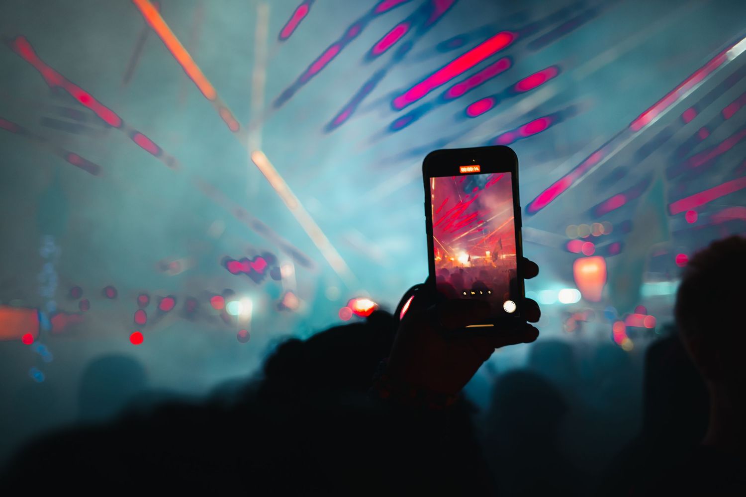 Silhouetted crowd holds up glowing smartphones at a concert with colorful lights streaming through smoke in the background.