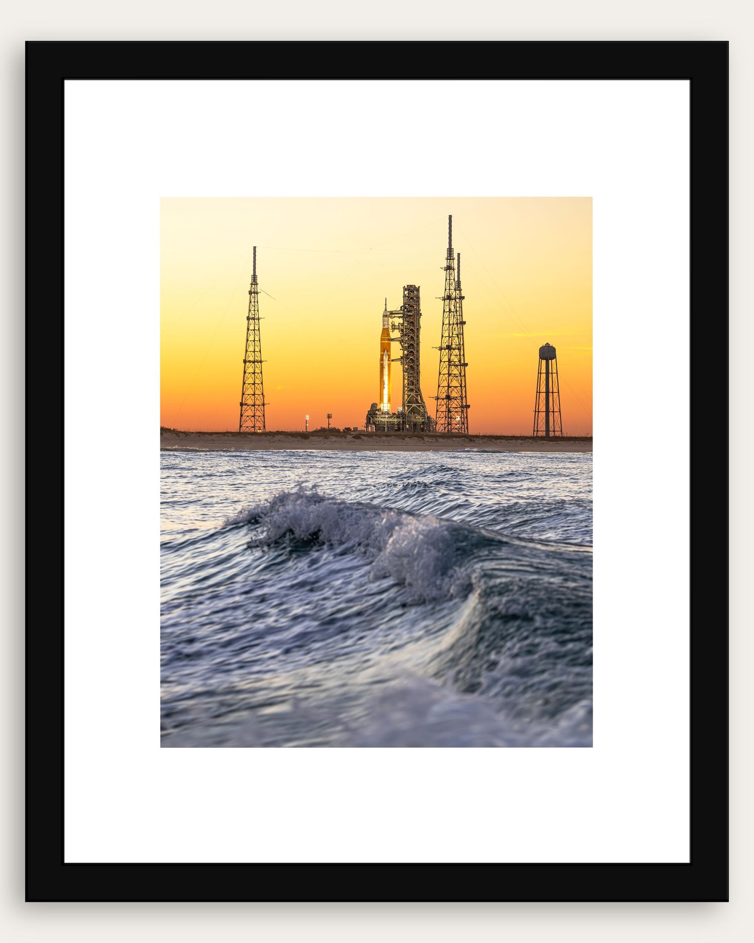 Framed photo of a rocket launch pad at sunset with ocean waves in the foreground, golden sky backdrop.