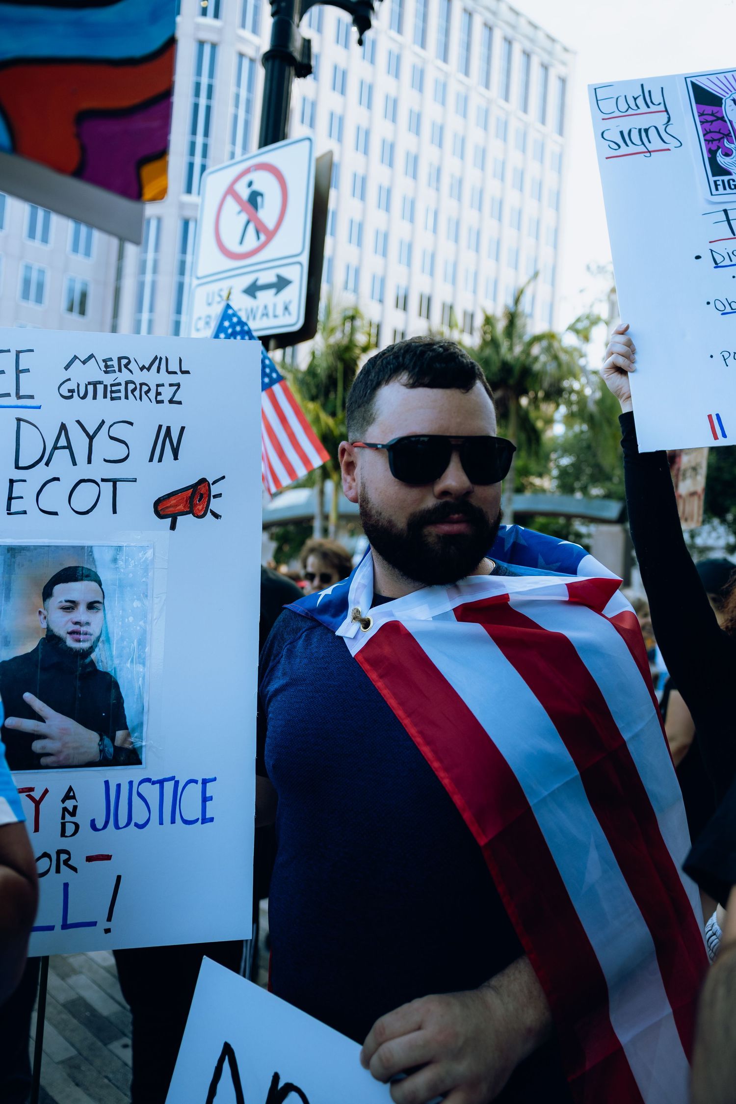 Demonstrators holding protest signs gather outside a government building with American flags visible.