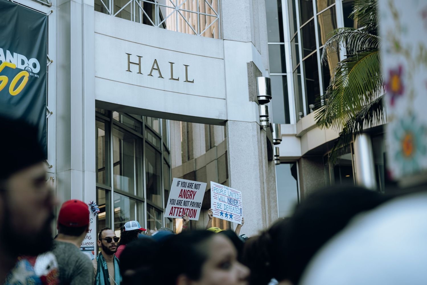 Protest crowd gathers outside a building labeled 'HALL' with visible signs and architectural details.