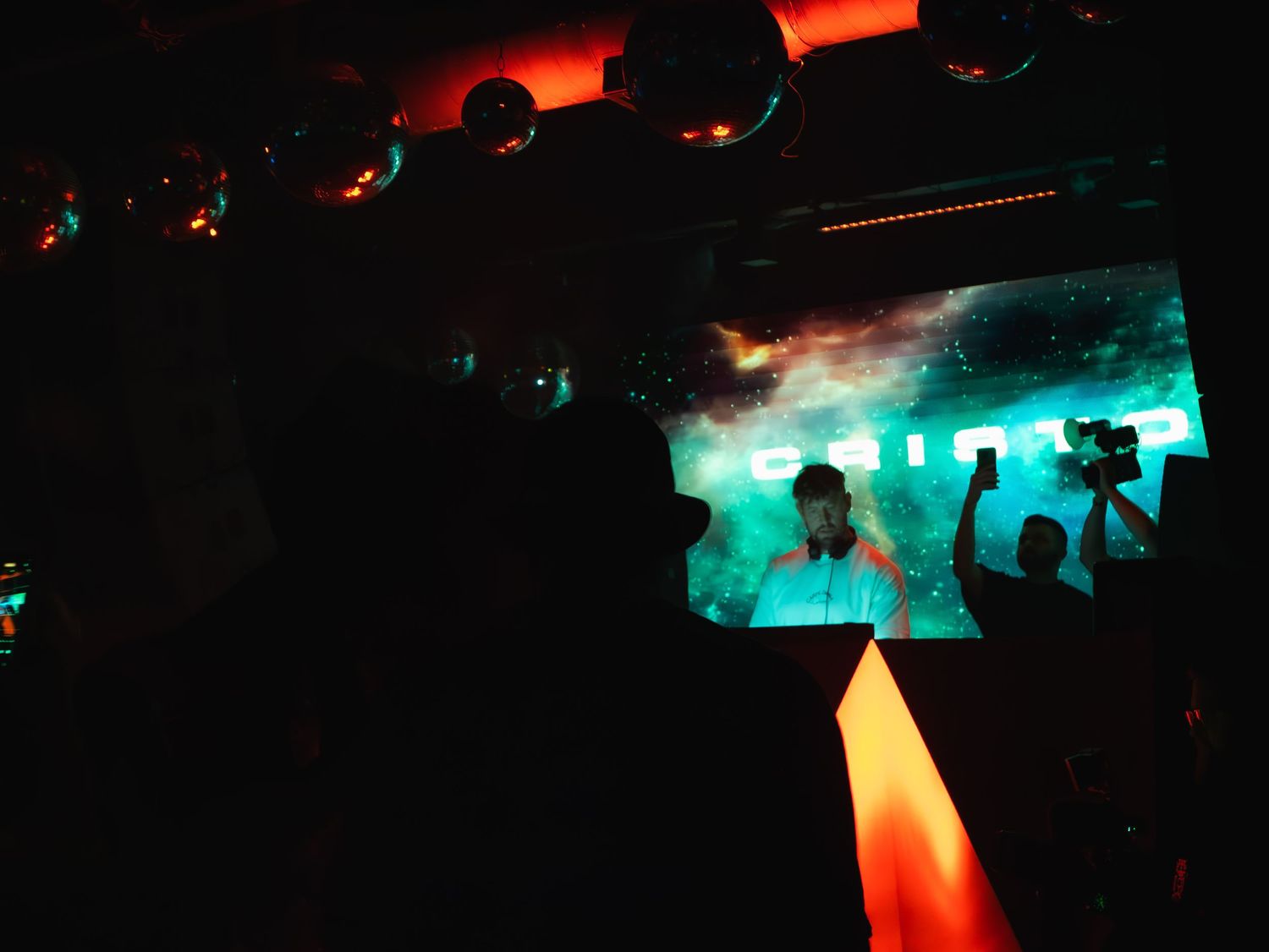 Silhouettes of concert crowd dancing beneath glowing turquoise and red lights in a dark nightclub atmosphere.