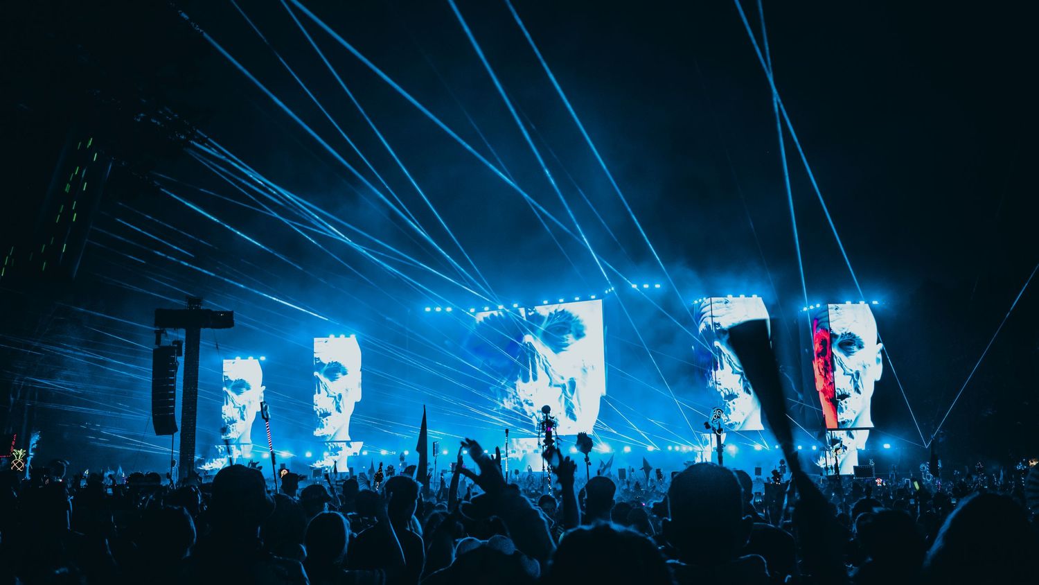 Concert crowd silhouettes raise their arms against a dramatic blue light show with beaming spotlights at a live performance.