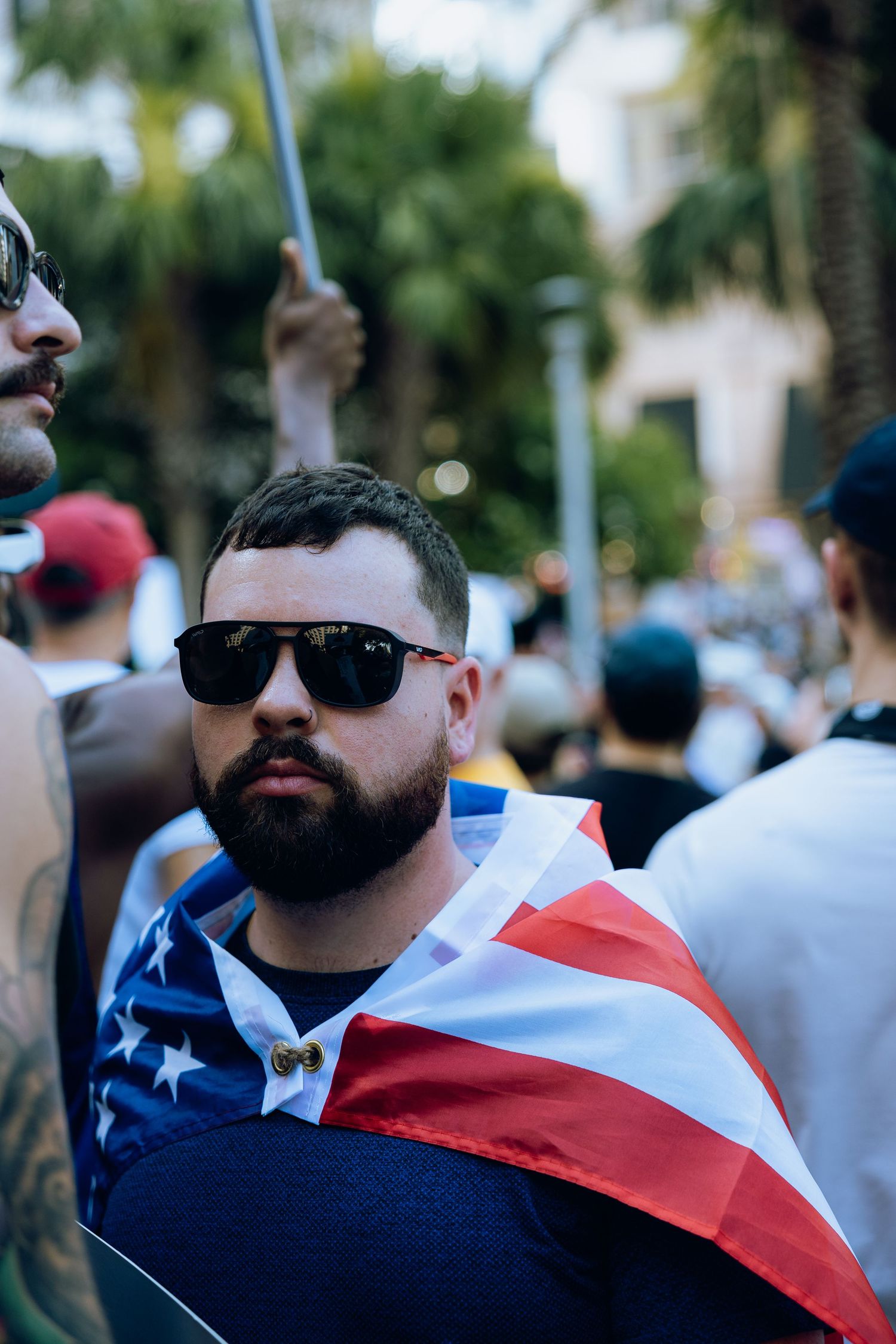 Protester wearing sunglasses and draped in American flag stands in crowd at political demonstration.
