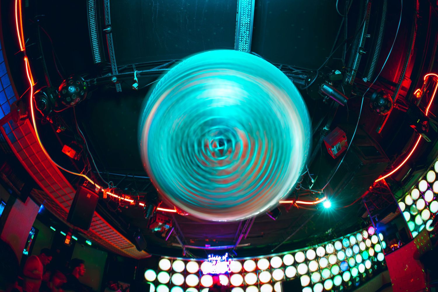 A glowing cyan spinning disc light installation on the ceiling of a vibrant nightclub with colorful LED lights.