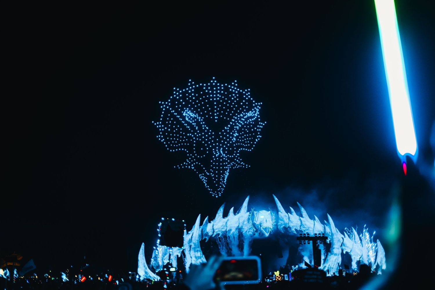 A drone light show forms a glowing phoenix shape above a blue-lit stage at an outdoor nighttime festival.