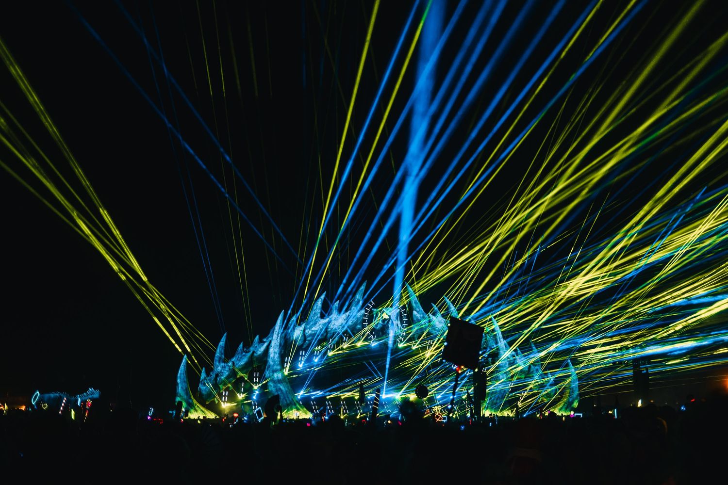 Colorful blue and yellow laser light beams illuminate a dark sky during an outdoor light show performance.